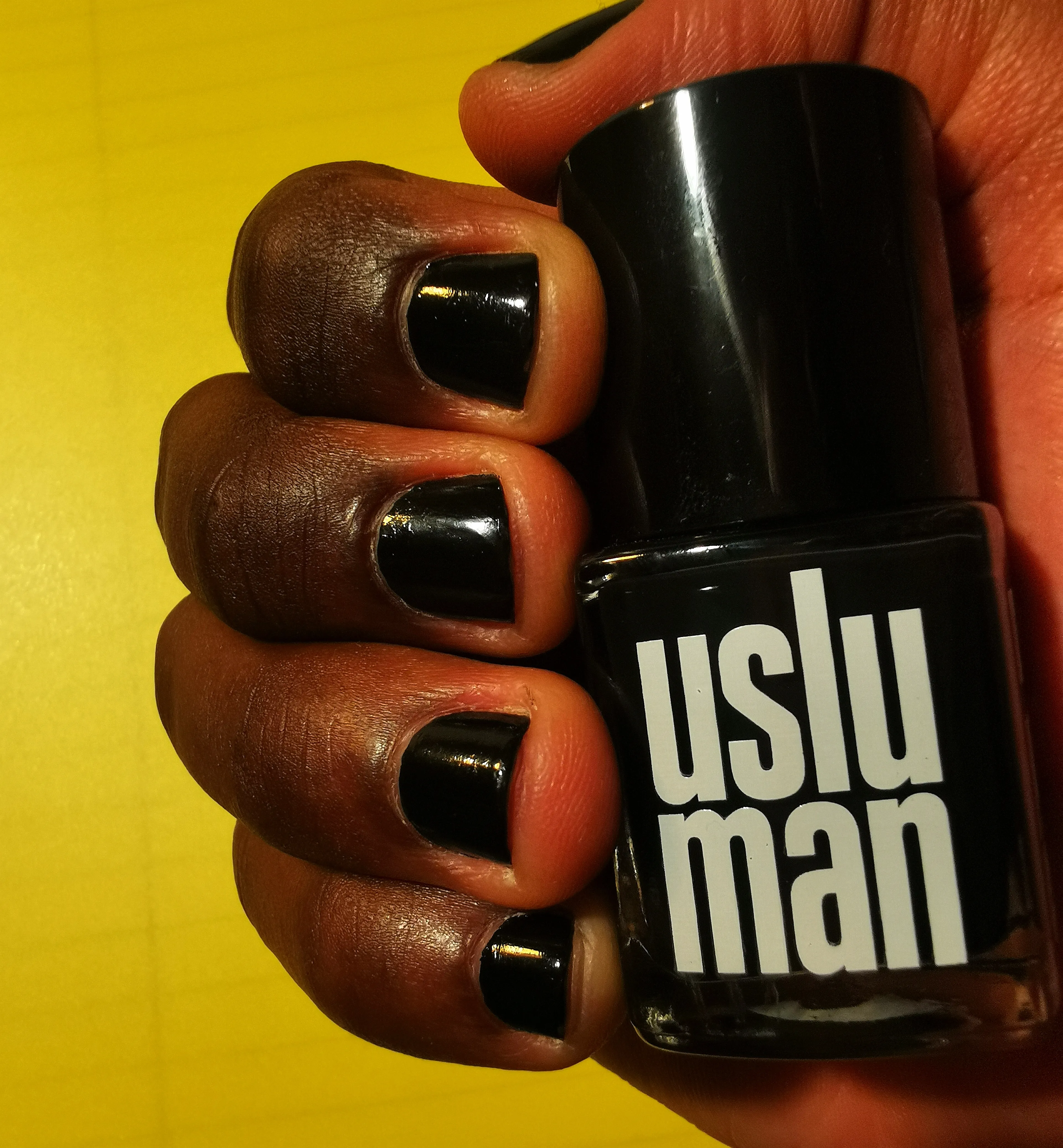 A shot of the Uslu Man Nail Polish in hand with fingers with glossy black nail polish, emphasizing the shine and smooth finish of the nail color against the warm skin tone and yellow background color.